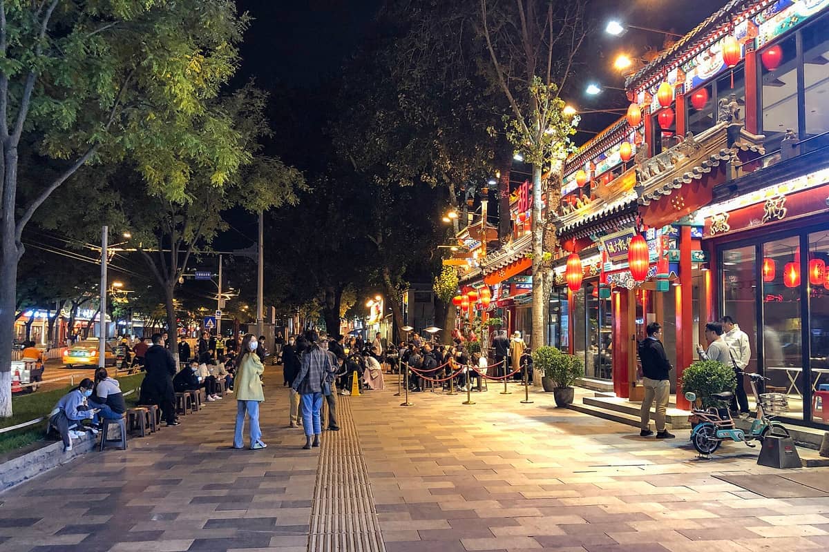 Guijie Ghost Street in Peking bei Nacht beleuchtet mit roten Laternen, belebten Restaurants und Menschengruppen auf dem Bürgersteig.