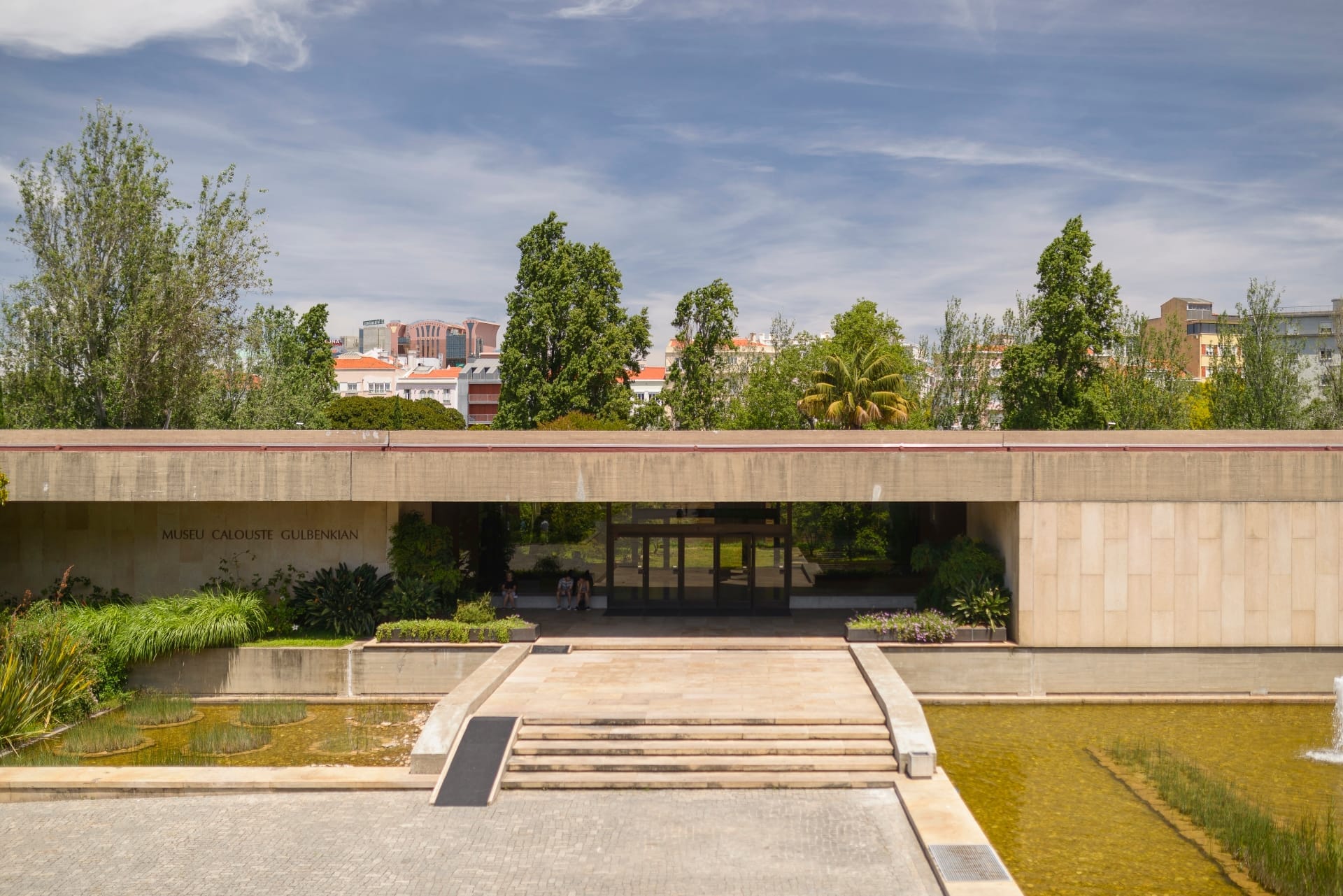 Ingresso del Museo Calouste Gulbenkian con facciata moderna in pietra, laghetto riflettente, rigogliosa vegetazione ed edifici della città sullo sfondo sotto un cielo azzurro.