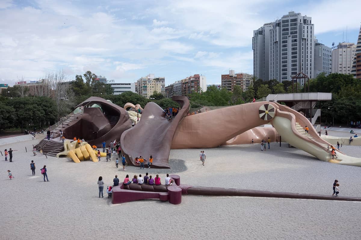 Grande scultura-parco giochi di Gulliver in vetroresina a Valencia, circondata da bambini che giocano e famiglie, con edifici e alberi della città sullo sfondo.