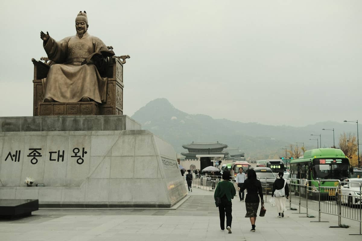 Vista ao nível da rua da Praça Gwanghwamun com a estátua do Rei Sejong, pedestres caminhando, ônibus verdes e montanhas ao fundo sob um céu nublado.