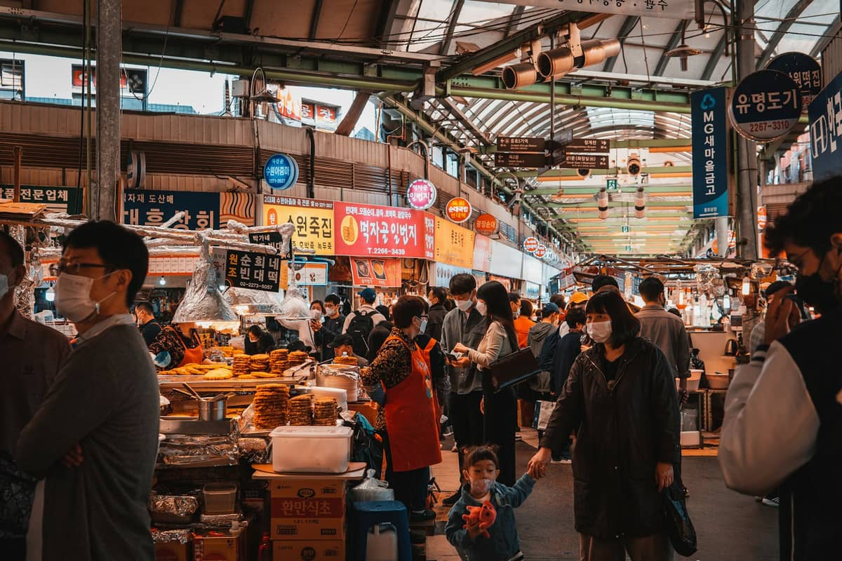 Uma movimentada viela gastronômica coberta dentro do Mercado Gwangjang em Seul, com muitas pessoas explorando as barracas sob um telhado iluminado e placas coloridas.
