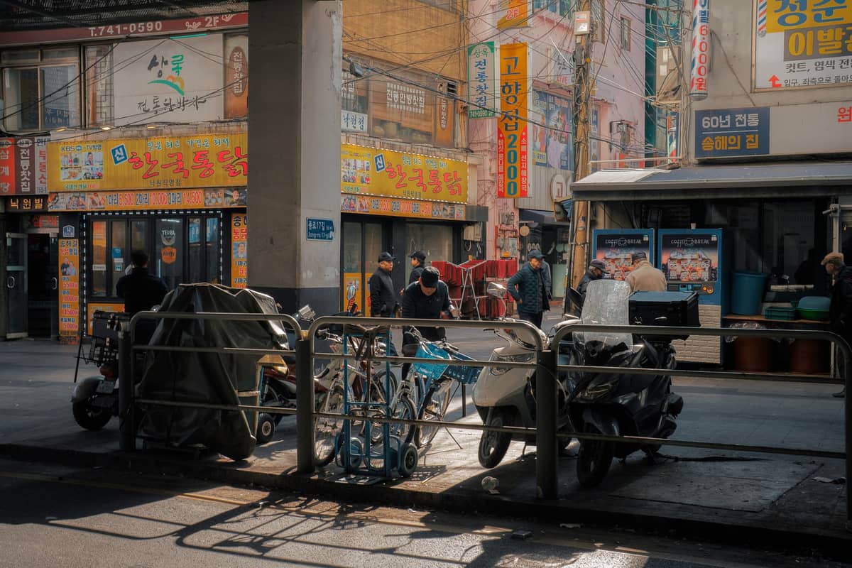 Vista diurna de uma rua perto do Mercado Gwangjang em Seul, com scooters, pessoas caminhando e lojas movimentadas com vibrantes letreiros em coreano.