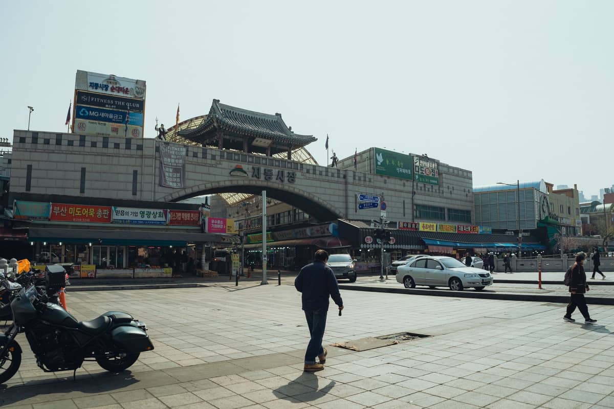 Vista ampla da entrada principal do Mercado Gwangjang em Seul, mostrando seu arco distinto e sinalização com pessoas e veículos na frente.