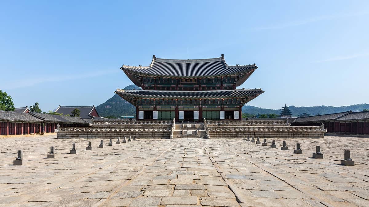 Vista panorâmica de Geunjeongjeon, o imponente salão do trono do Palácio Gyeongbokgung, com um pátio de pedra e montanhas ao fundo sob um céu azul e limpo.