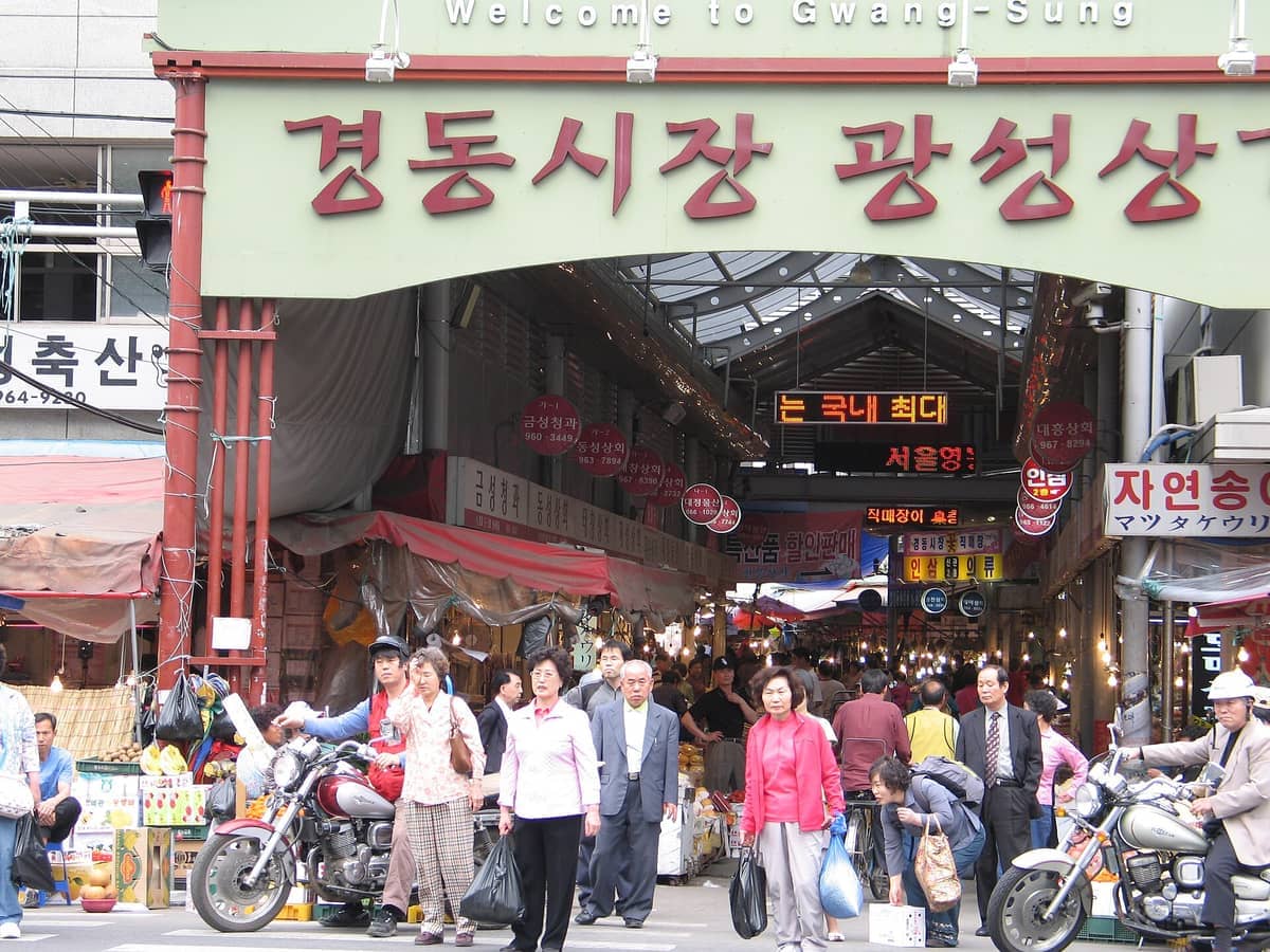 Entrada do Mercado Gyeongdong em Seul, movimentada com pessoas, motocicletas e barracas vibrantes vendendo frutas e produtos tradicionais sob uma passagem coberta.