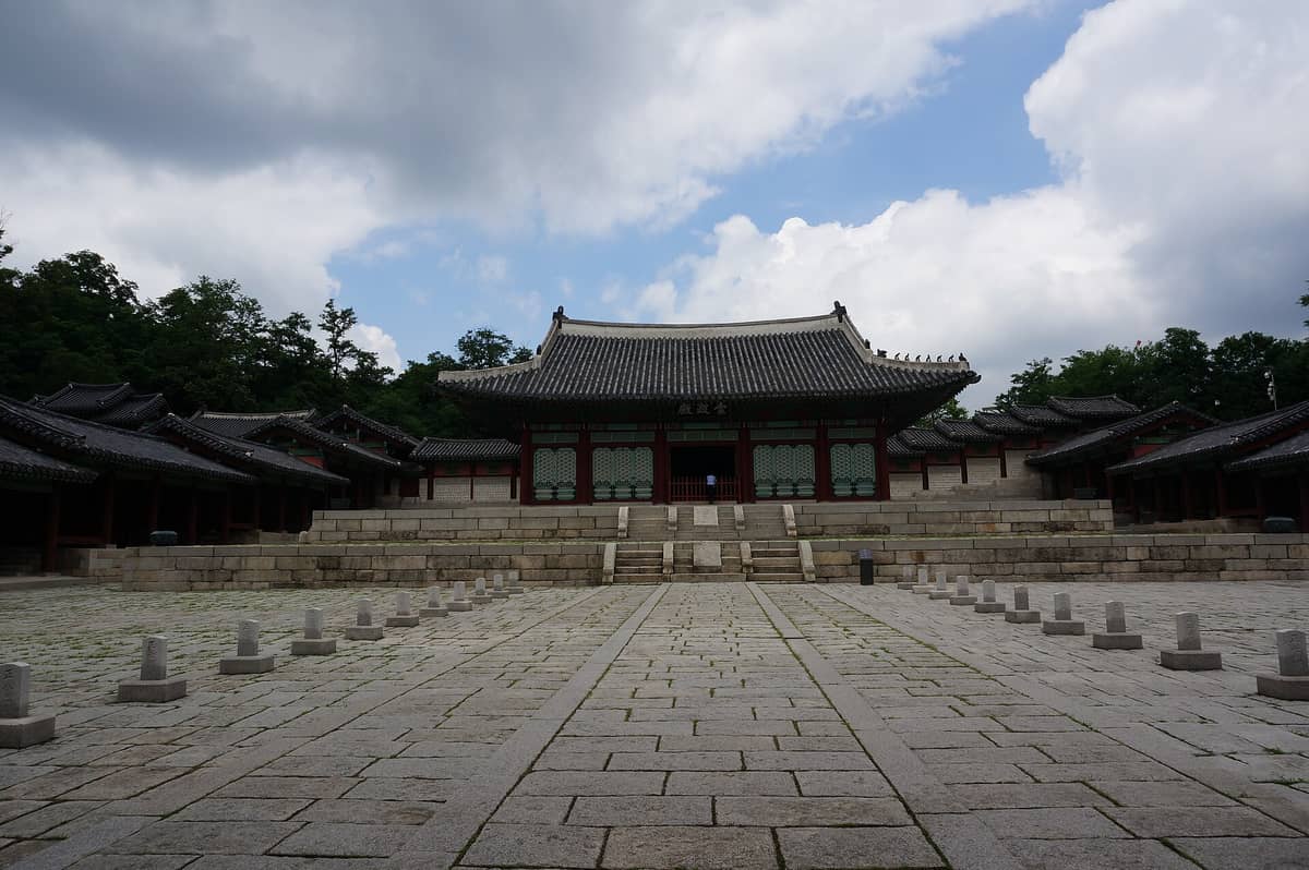 Vista ampla do salão principal do Palácio Gyeonghuigung com sua plataforma de pedra elevada, ladeado por construções tradicionais, cercado de árvores, sob um céu parcialmente nublado em Seul.