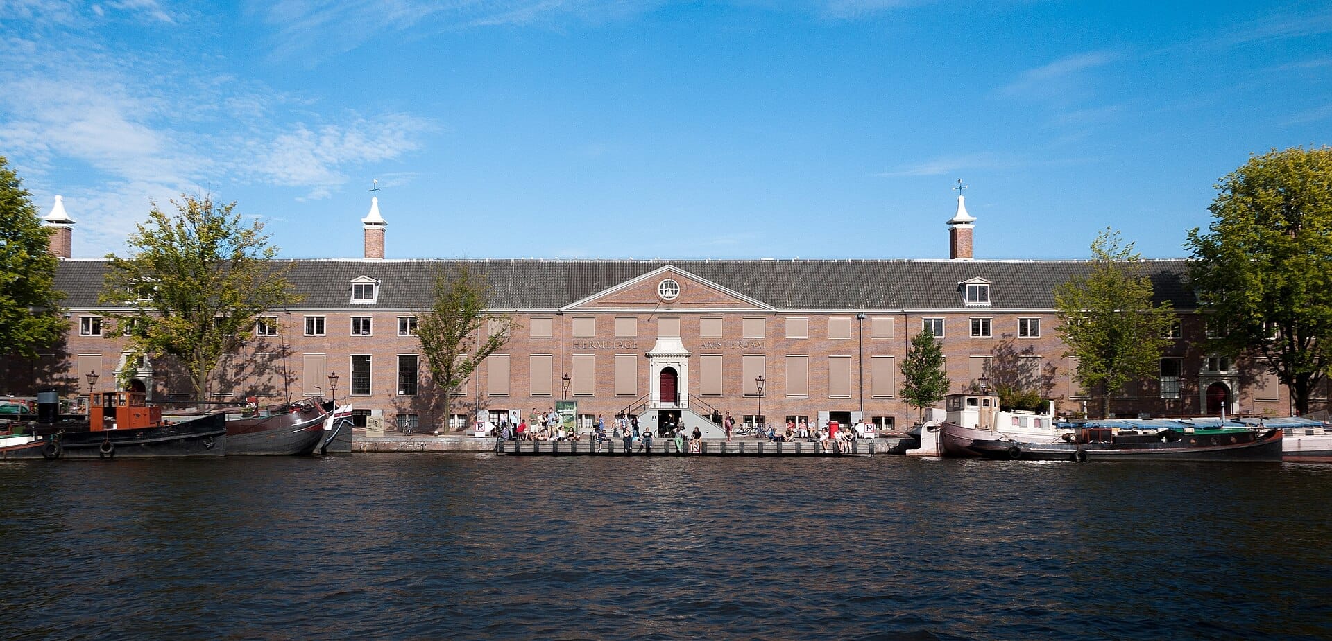 El H'ART Museum Amsterdam visto desde el otro lado del río Amstel, con su histórica fachada de ladrillo, barcas amarradas en frente y visitantes entrando al museo.