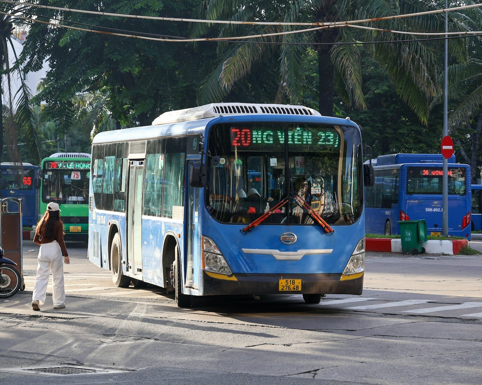 Blue public city bus in front of palm trees at a Hanoi intersection, with street activity and clear signage.