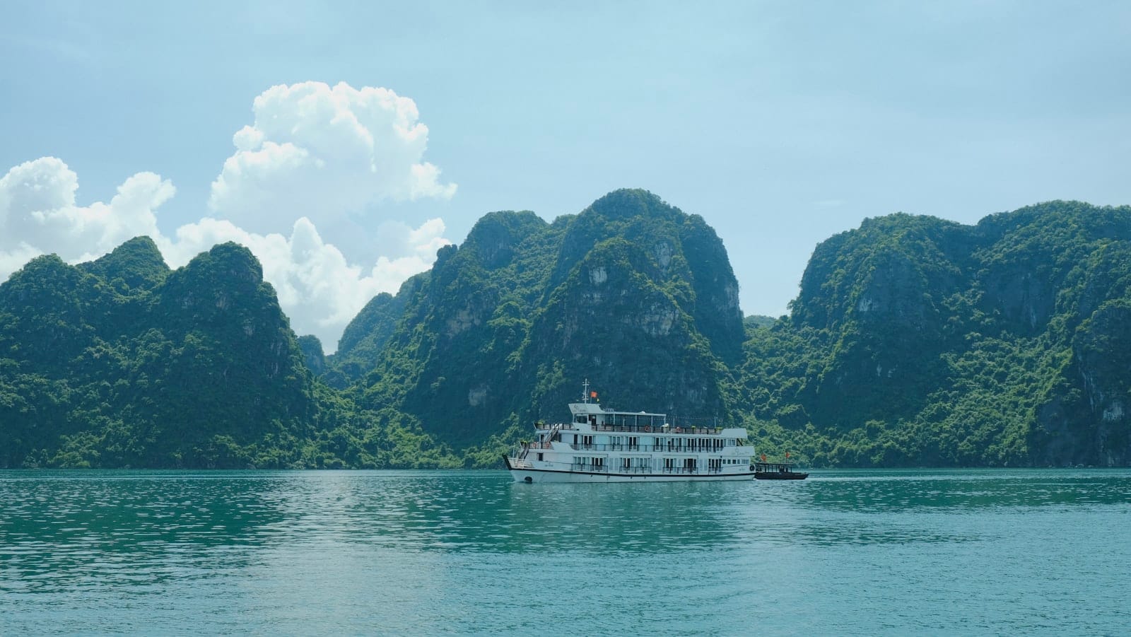 A large white cruise boat sails through emerald waters surrounded by dramatic limestone karsts under a bright blue sky in Ha Long Bay, Vietnam.