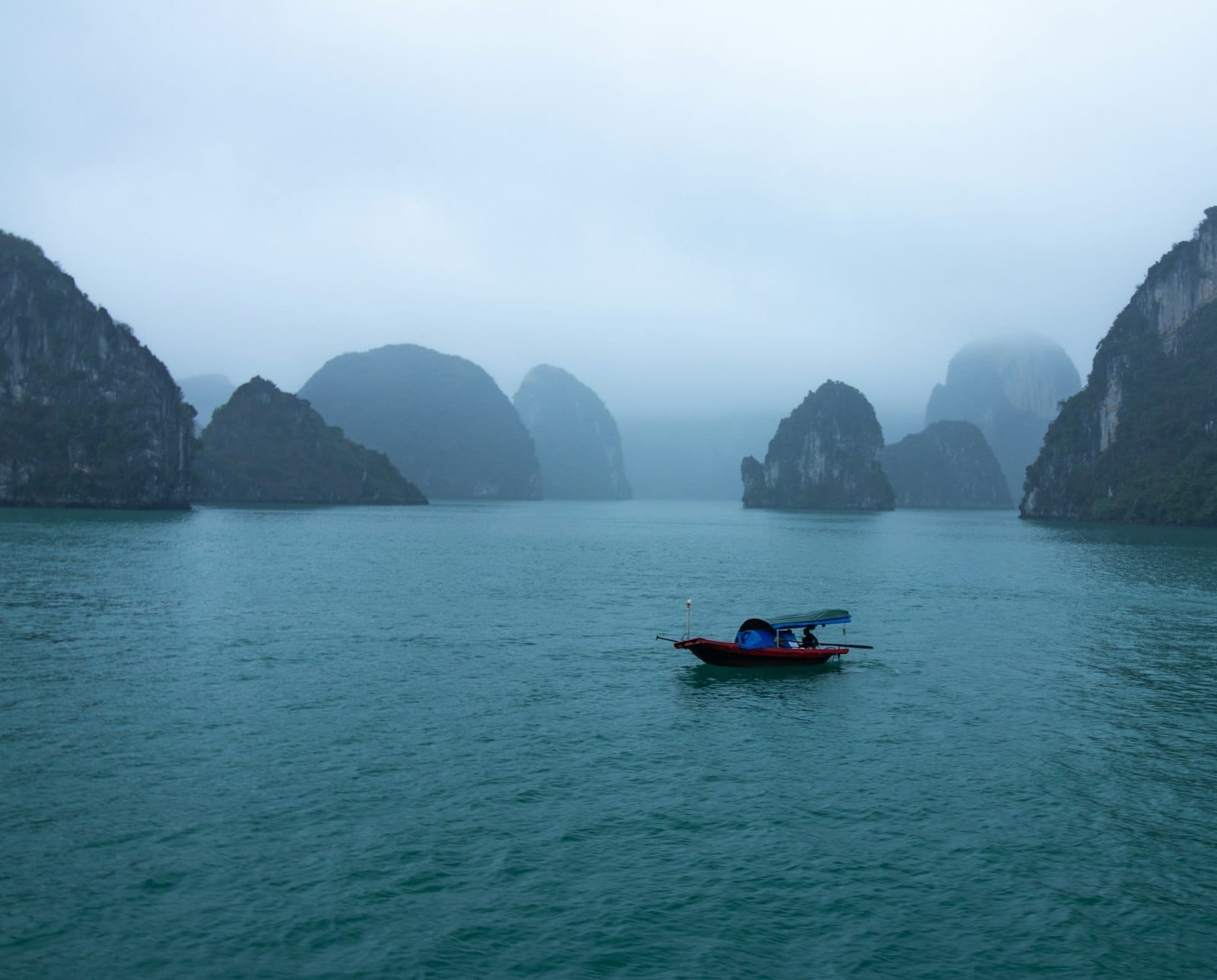 Small boat on turquoise water with limestone karsts in background and misty sky at Ha Long Bay.