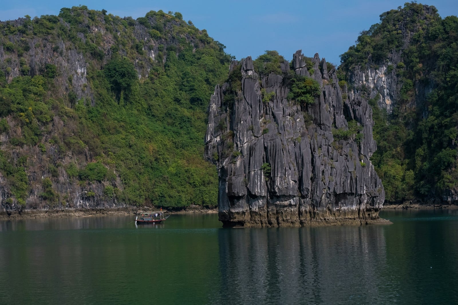 Tall limestone karst rock formation surrounded by calm green water and lush hills, with a small wooden boat nearby.