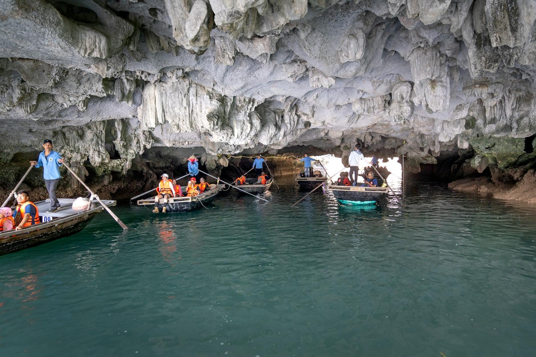 Tourists in rowboats wearing life vests passing through a limestone cave on turquoise water in Ha Long Bay.