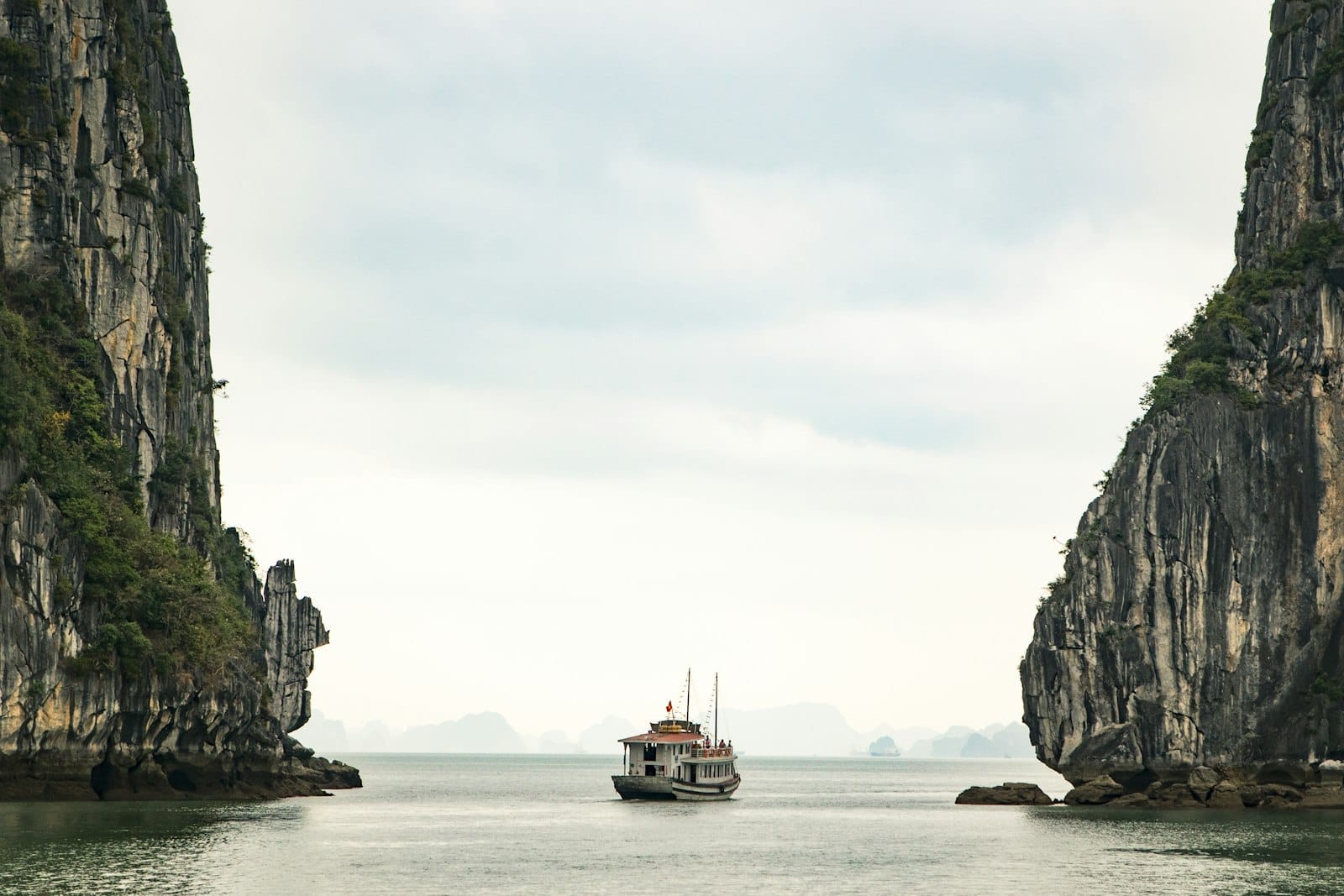 A scenic cruise boat sails between towering limestone cliffs in Ha Long Bay under a cloudy sky, showcasing dramatic seascape and iconic karst formations.