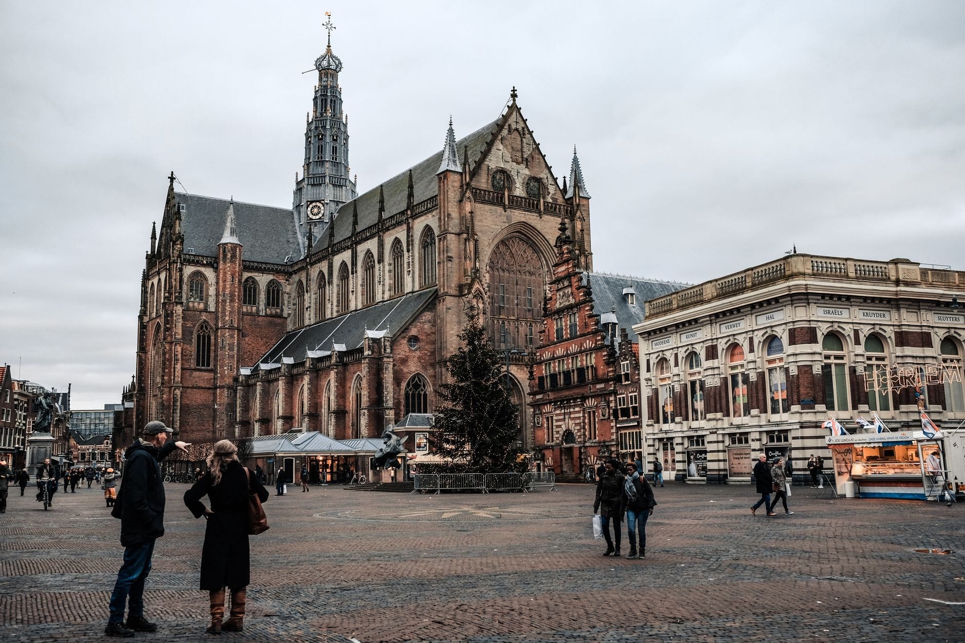 Toàn cảnh nhà thờ Grote Kerk và quảng trường chợ sôi động của Haarlem với nhiều người qua lại, nhà thờ lịch sử uy nghi hiện ra dưới bầu trời nhiều mây.
