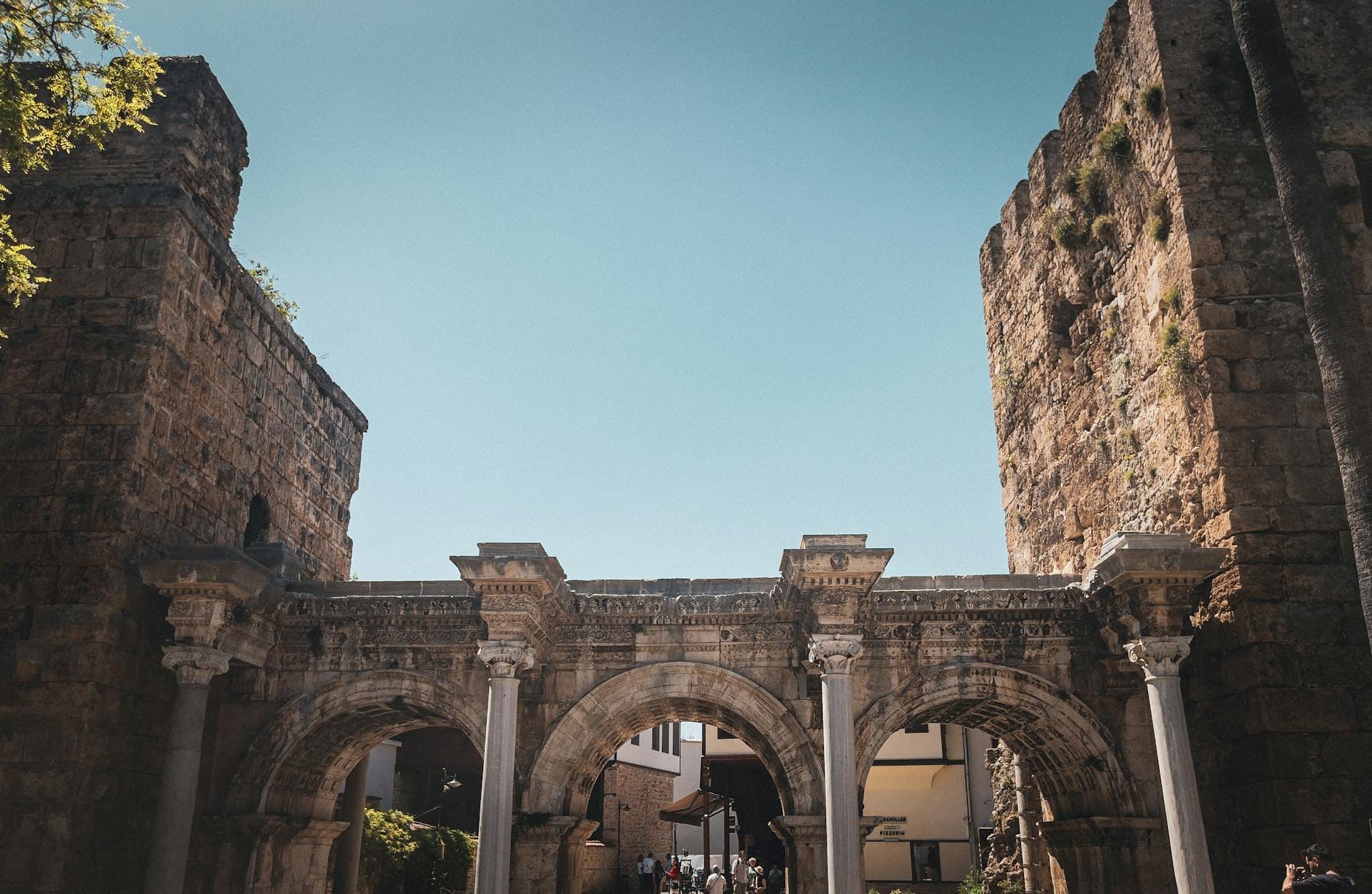 Vista grandangolare della Porta di Adriano ad Antalya con i tre archi romani in marmo incorniciati da antiche torri in pietra sotto un cielo azzurro.