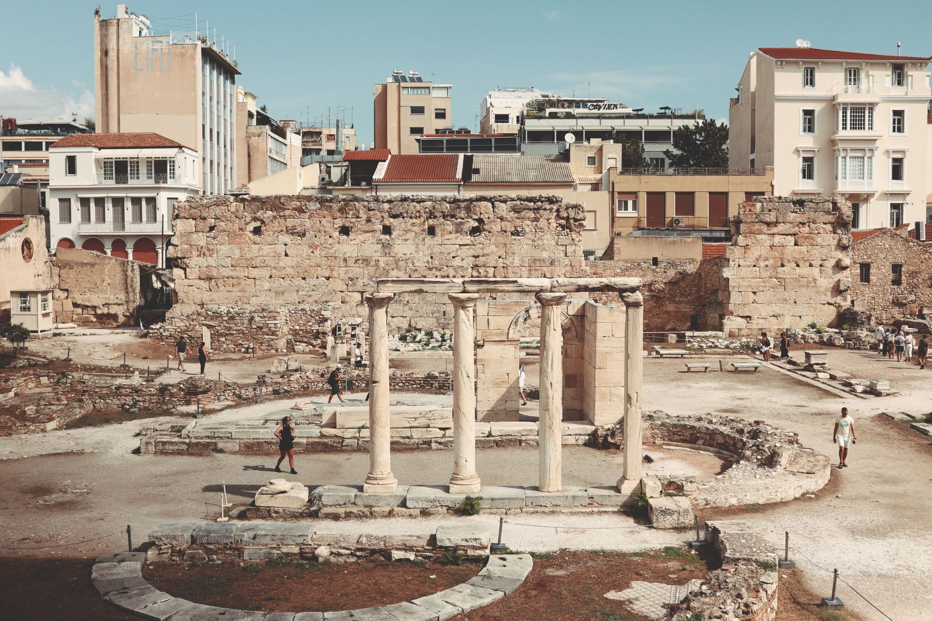 Vista ampia delle colonne in rovina e delle fondamenta della Biblioteca di Adriano, con antiche mura in pietra e gli edifici moderni di Atene sullo sfondo sotto un cielo terso.