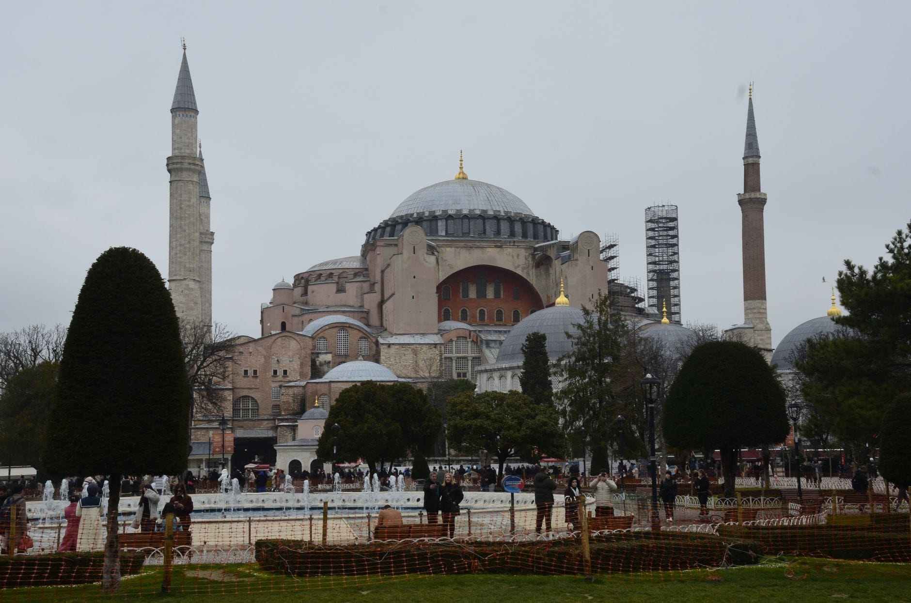 Vista grandangolare dell'esterno di Hagia Sophia con cupole e minareti, circondata da alberi, giardini e turisti sotto un cielo nuvoloso.
