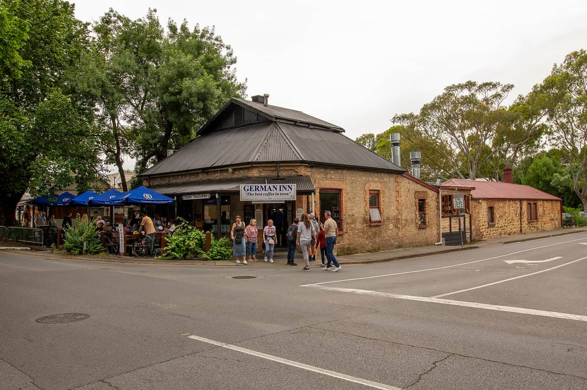 The German Inn in Hahndorf, a stone heritage building with blue umbrellas and people gathered in front, surrounded by leafy trees.