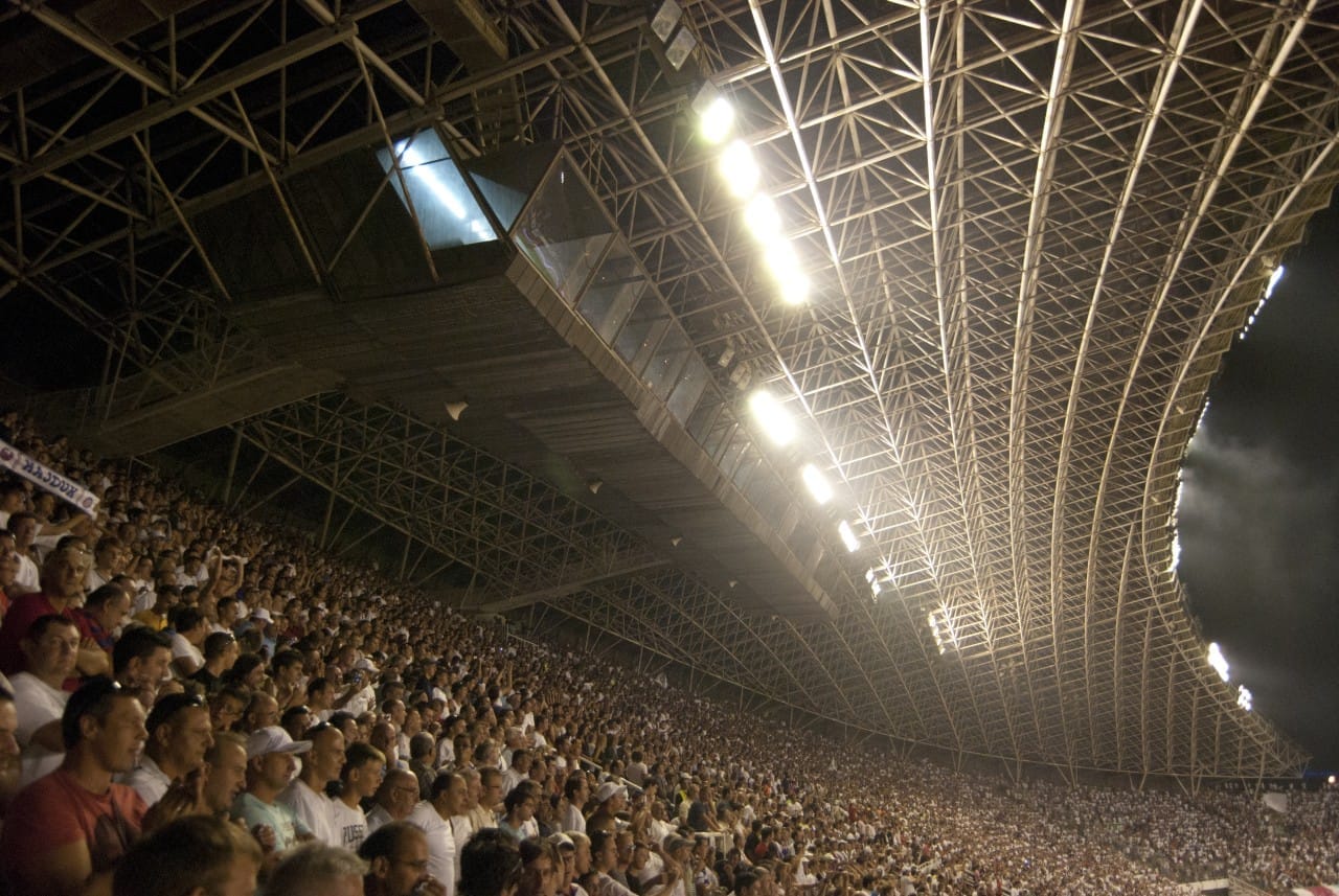 Arquibancadas lotadas no Estádio Poljud, Split, repletas de torcedores de futebol sob o distinto telhado curvo de aço e as luzes brilhantes do estádio.
