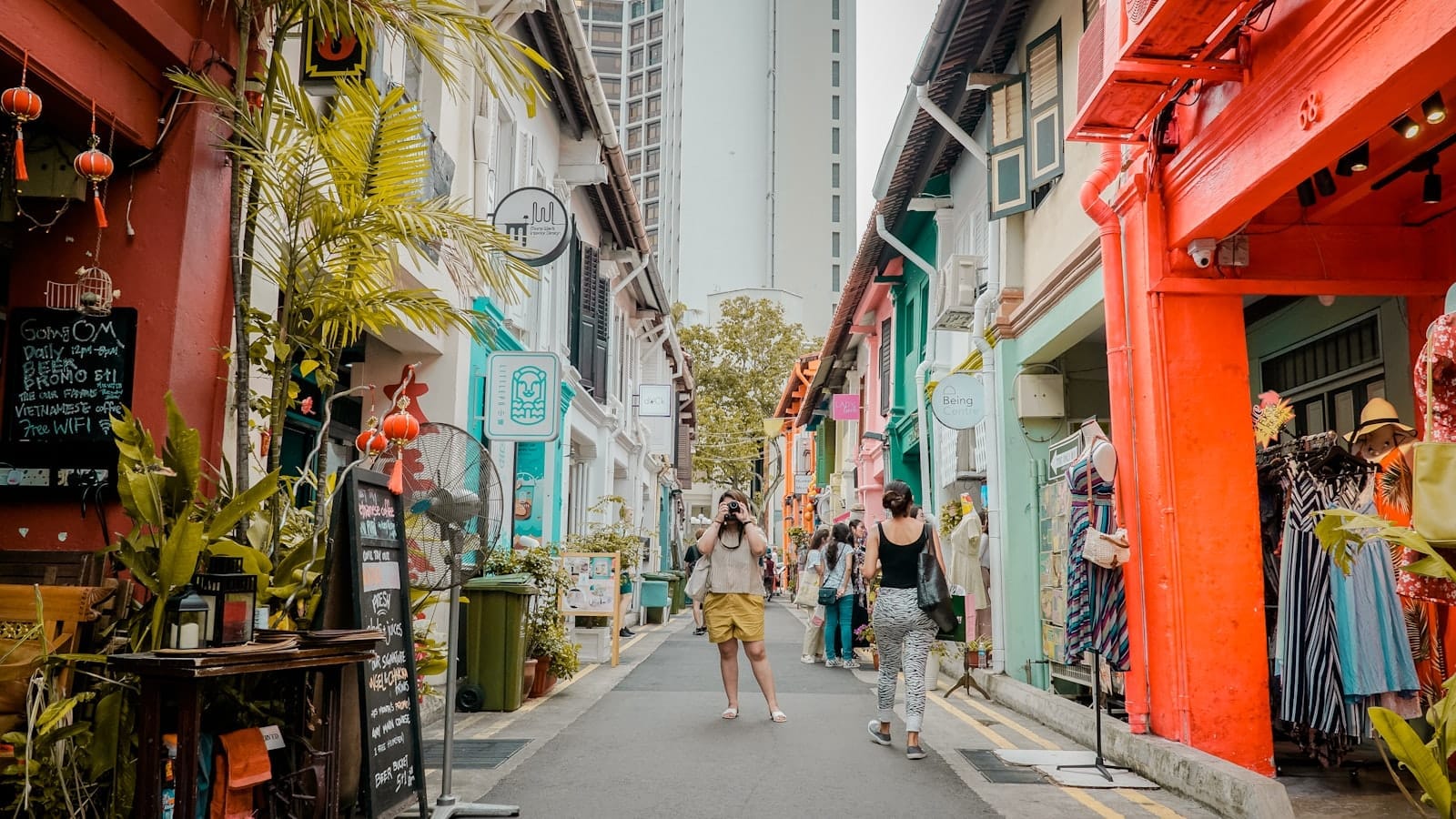 ถนนแคบๆ สีสันสดใสของฮาจิ เลน เรียงรายด้วยตึกแถวสีพาสเทล หน้าร้านสดใส ต้นไม้ริมทาง และผู้คนเดินเที่ยวท่ามกลางแสงกลางวัน