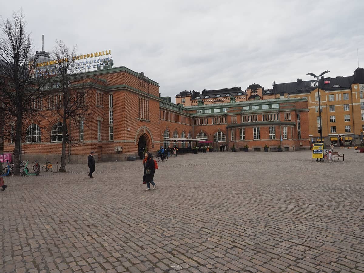 O Mercado de Hakaniemi em Helsinki visto de uma praça de paralelepípedos, com pessoas caminhando em frente à sua histórica fachada de tijolos vermelhos em um dia nublado.