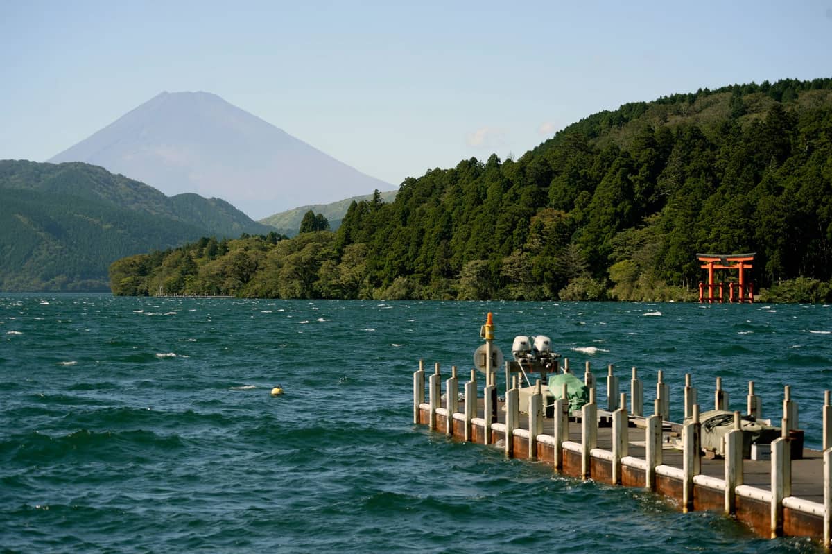 Hakone’s Lake Ashi with a view of Mount Fuji in the background and a red torii gate on the shore, seen from a wooden pier jutting into the water.