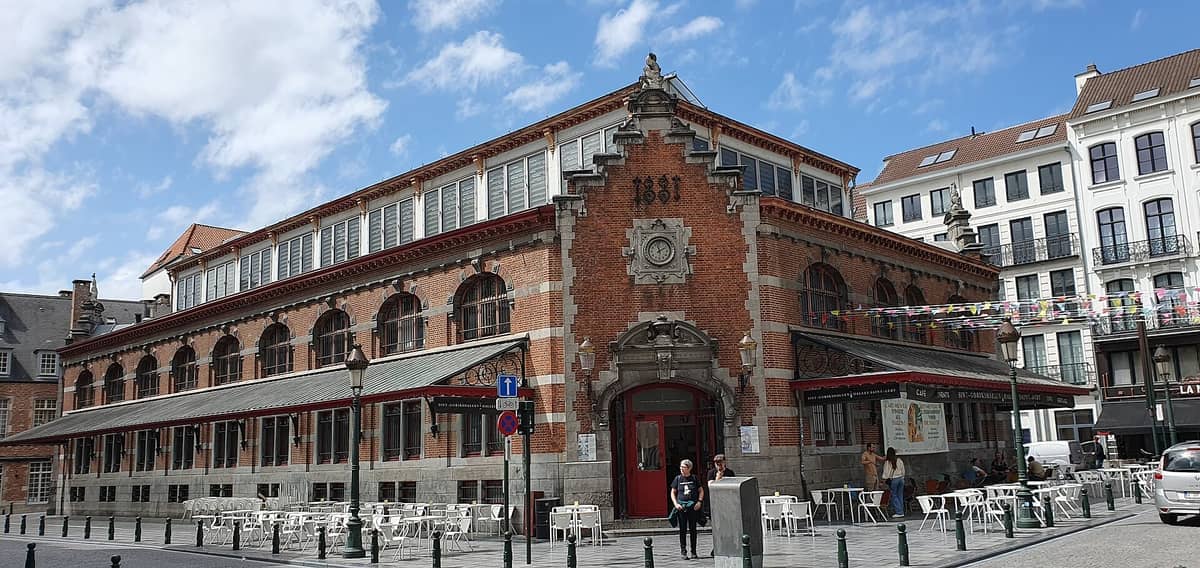 Vista exterior das Halles Saint-Géry, um histórico mercado de tijolos vermelhos com janelas em arco, mesas de café na rua e céu azul.