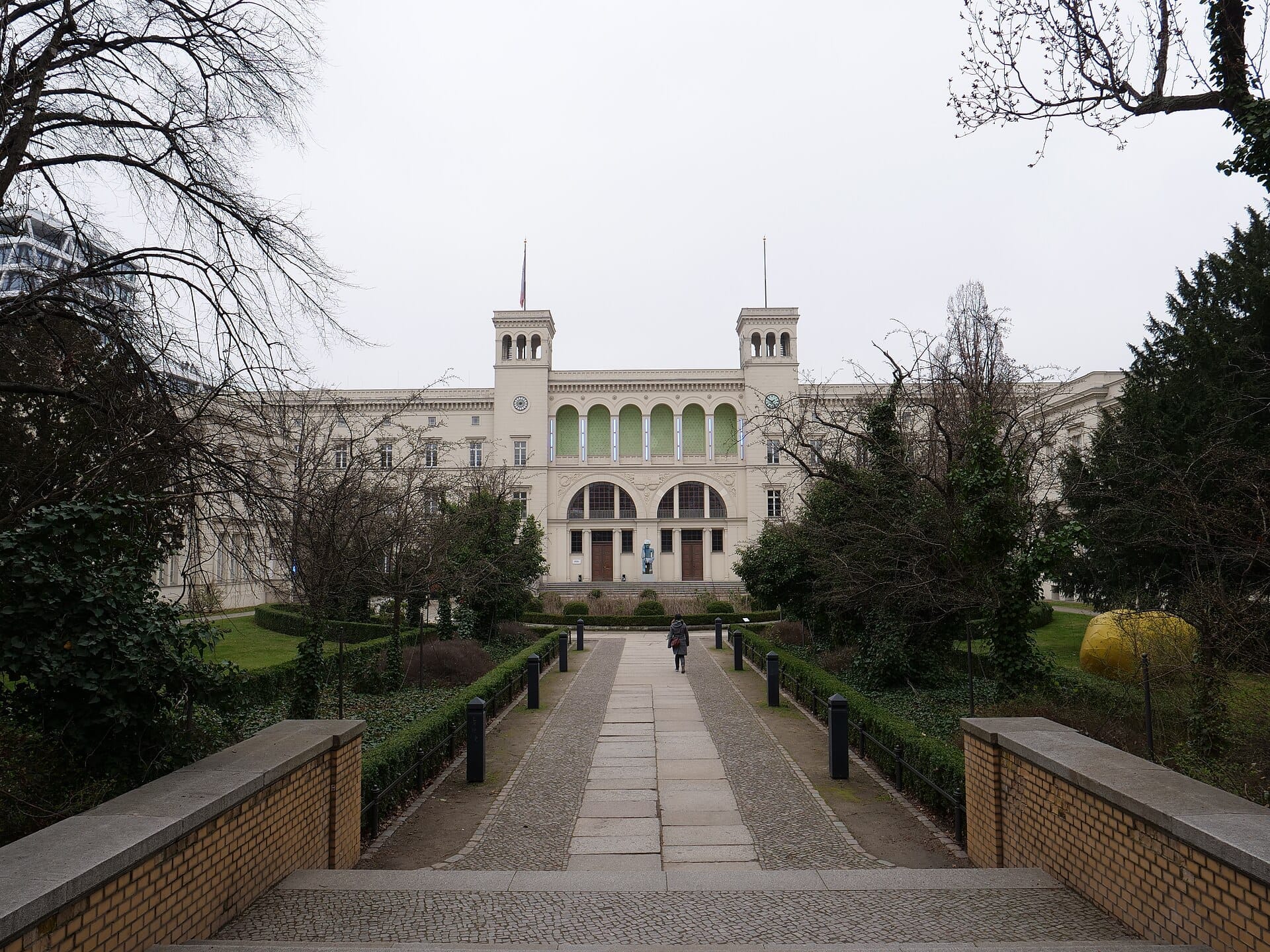 Hamburger Bahnhof a Berlino, un grandioso edificio neoclassico con due torri, visto da un vialetto paesaggistico in una giornata grigia e nuvolosa.