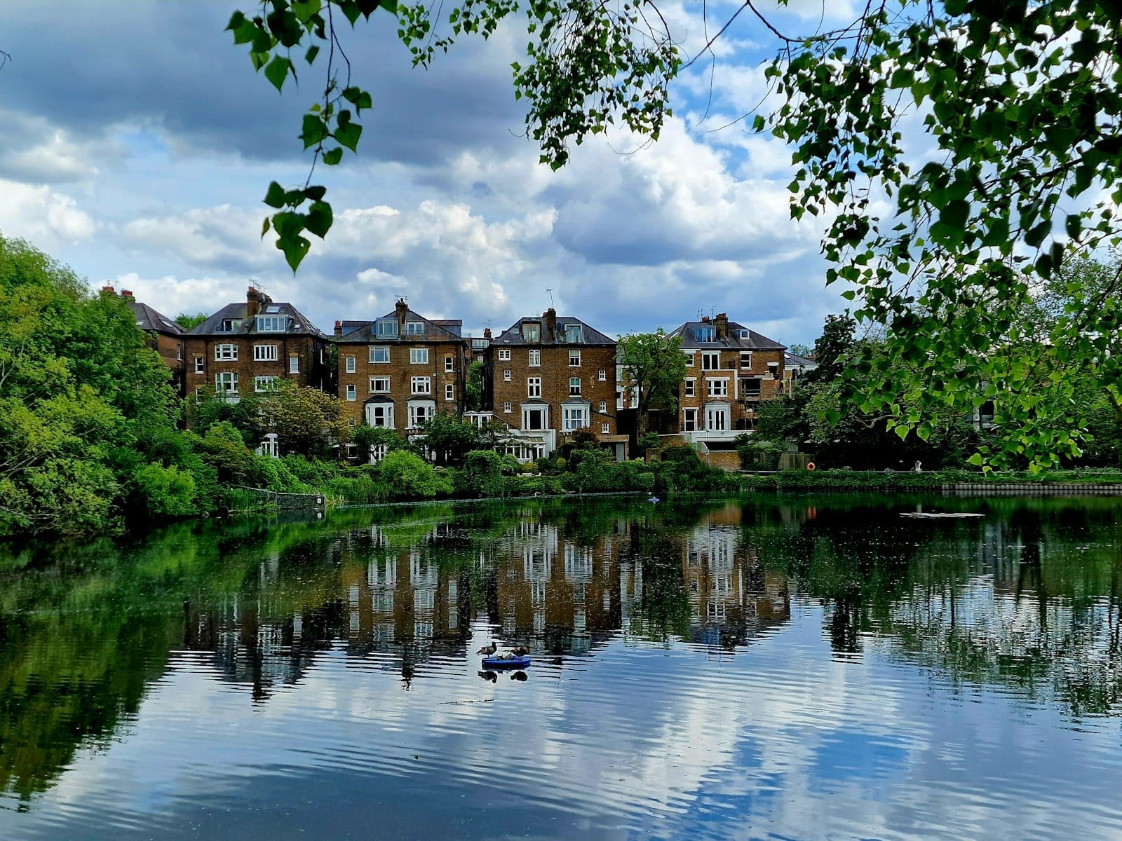 บ้านอิฐสวยริมบ่อน้ำใน Hampstead Heath สะท้อนเงาอยู่กลางต้นไม้เขียวชอุ่มและท้องฟ้ามีเมฆครึ้ม บรรยากาศอบอุ่นของแฮมป์สเตด & ไฮเกต
