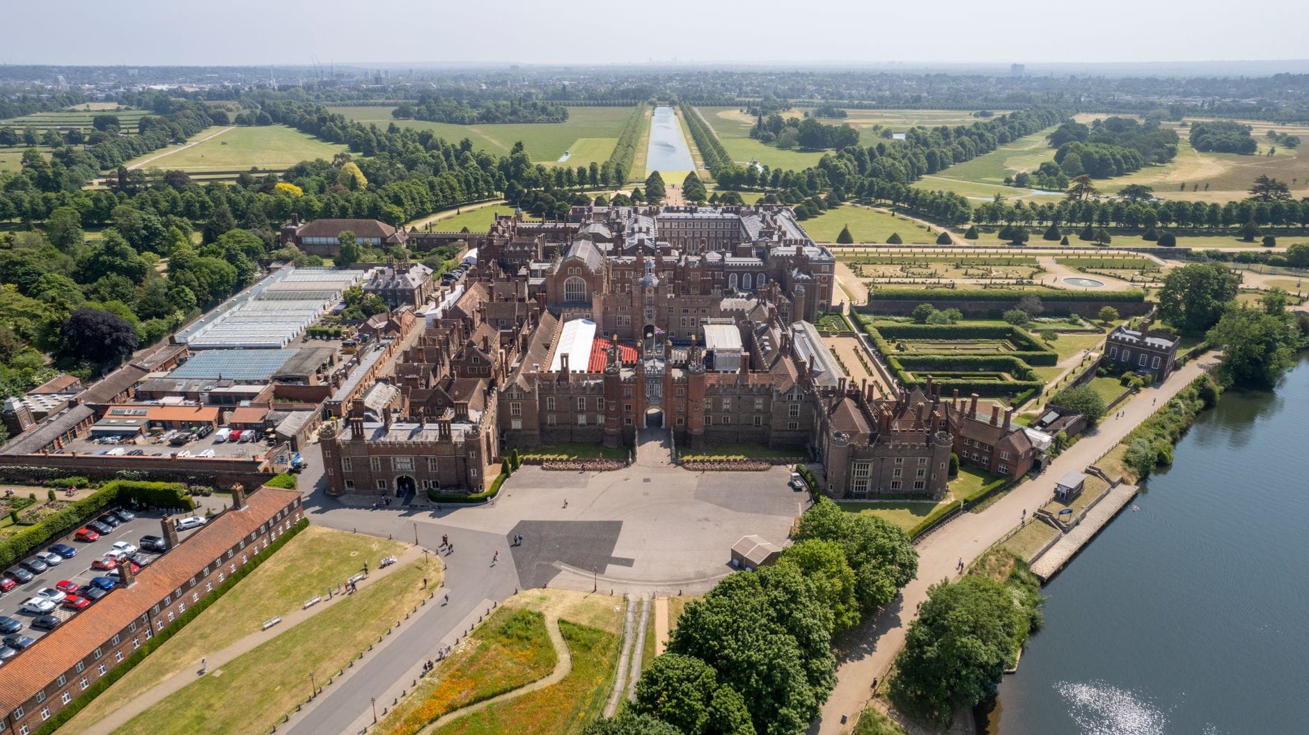 Una vista aerea mozzafiato di Hampton Court Palace, circondata da rigogliosi giardini, il Tamigi e un'ampia distesa di parco sotto un cielo terso.