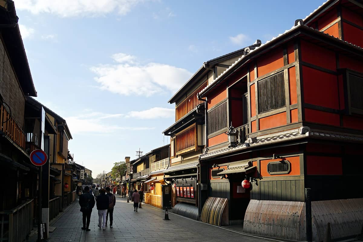 Hanamikoji Street in Kyoto’s Gion district with traditional wooden machiya buildings, ochaya teahouses, and a few pedestrians walking under a bright blue sky.