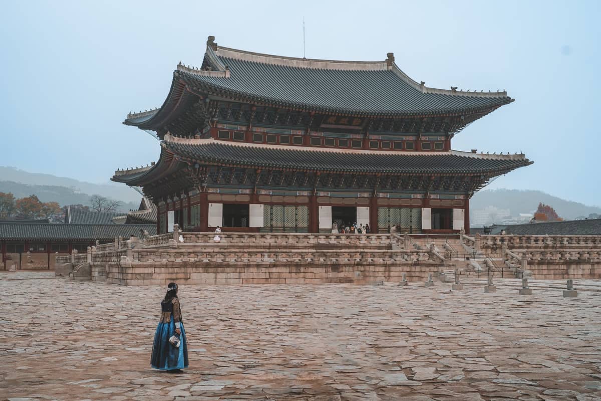 Uma pessoa vestindo um hanbok tradicional em frente ao hall principal do Palácio Gyeongbokgung, em Seul, Coreia do Sul, num pátio de pedra.
