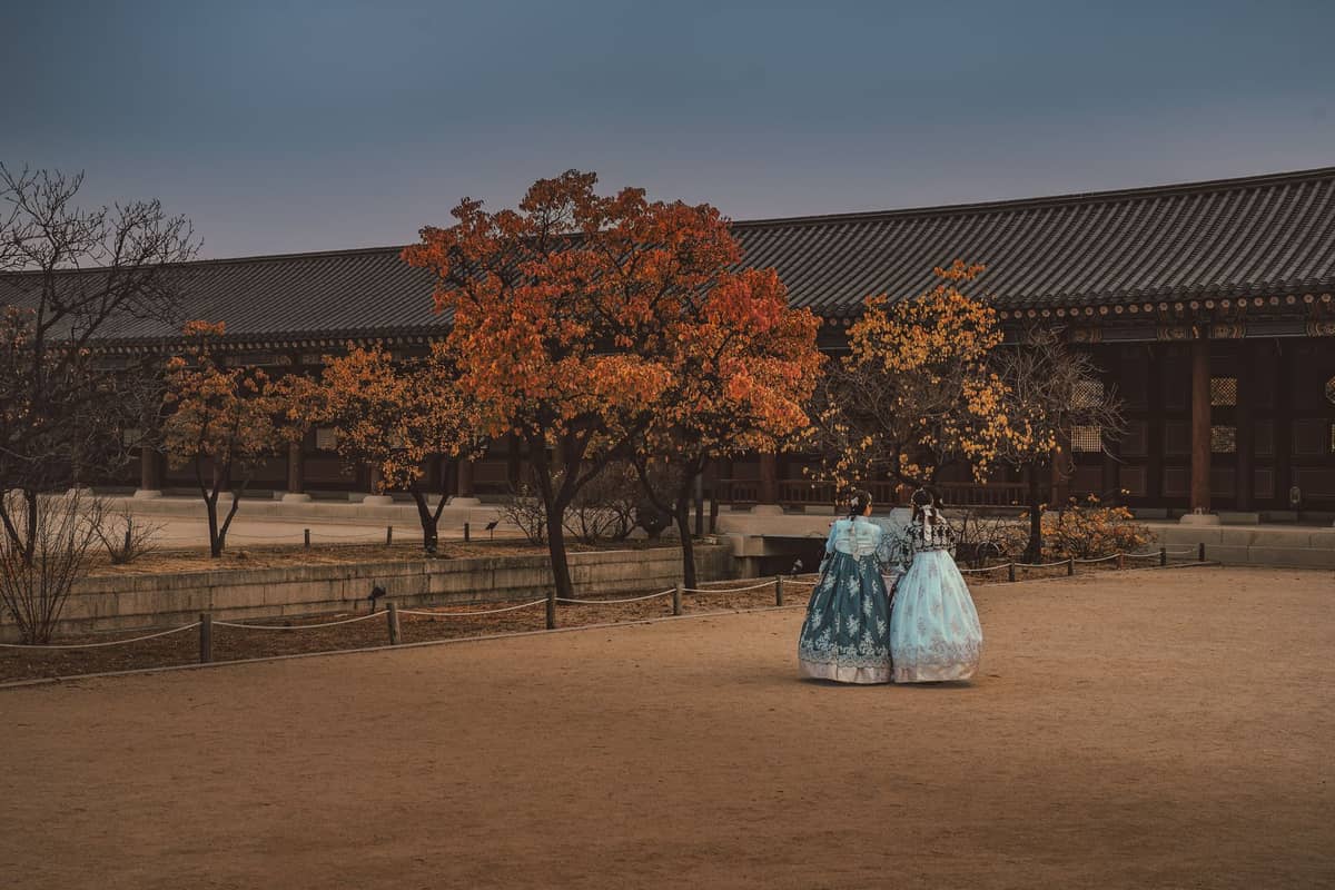 Duas pessoas com hanbok tradicional caminham perto de uma muralha de palácio rodeada de folhagem outonal colorida em Seul.
