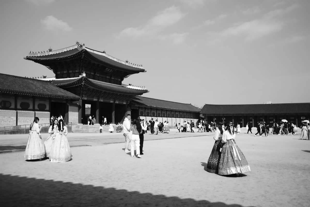 Visitantes com hanbok e roupas modernas entram por um grande portão de palácio em Seul, com o pátio do palácio movimentado em primeiro plano.