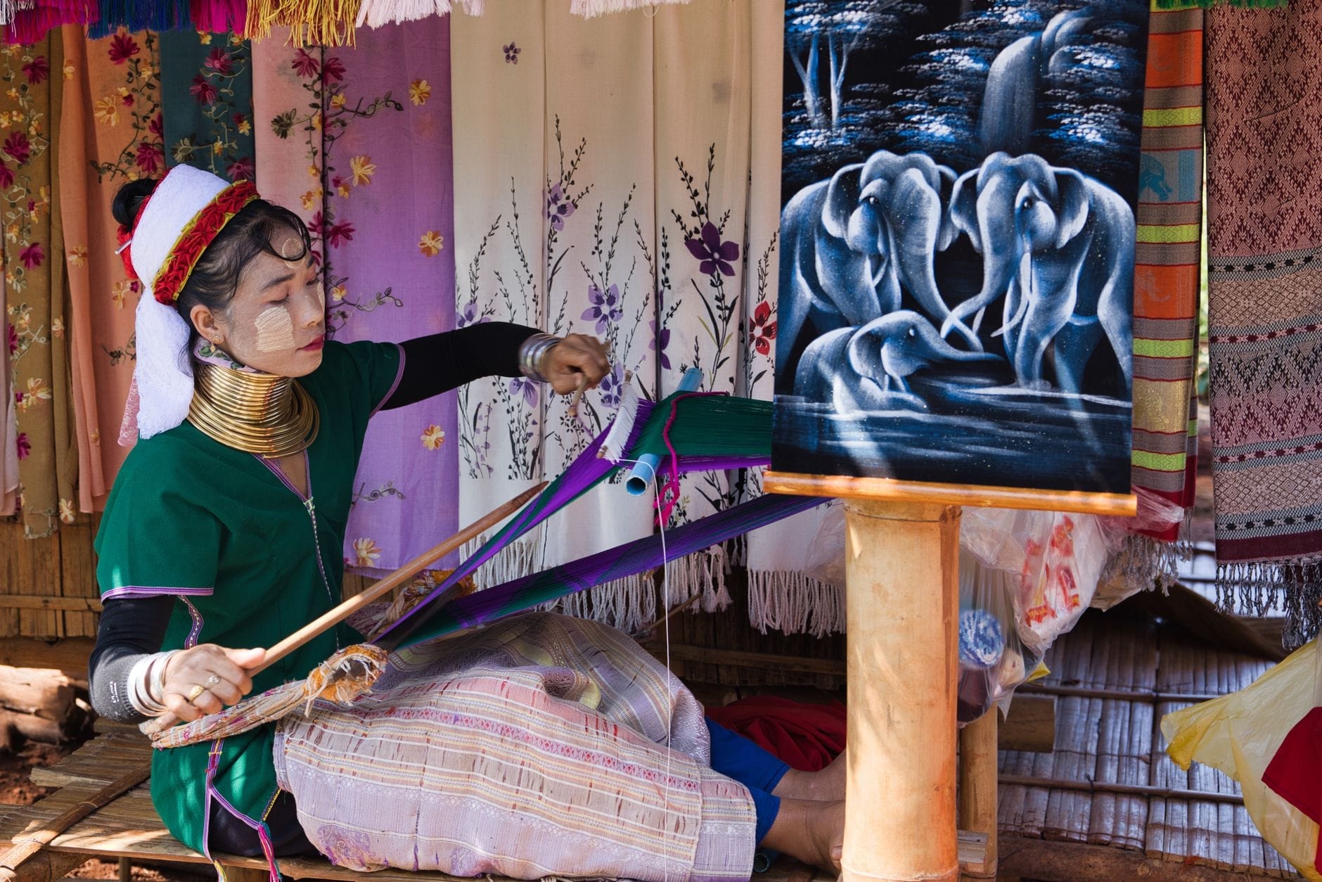 Una mujer con vestimenta tradicional tejiendo hábilmente en un telar, rodeada de coloridas telas bordadas y un intrincado tapiz de elefantes en un entorno de pueblo artesanal.