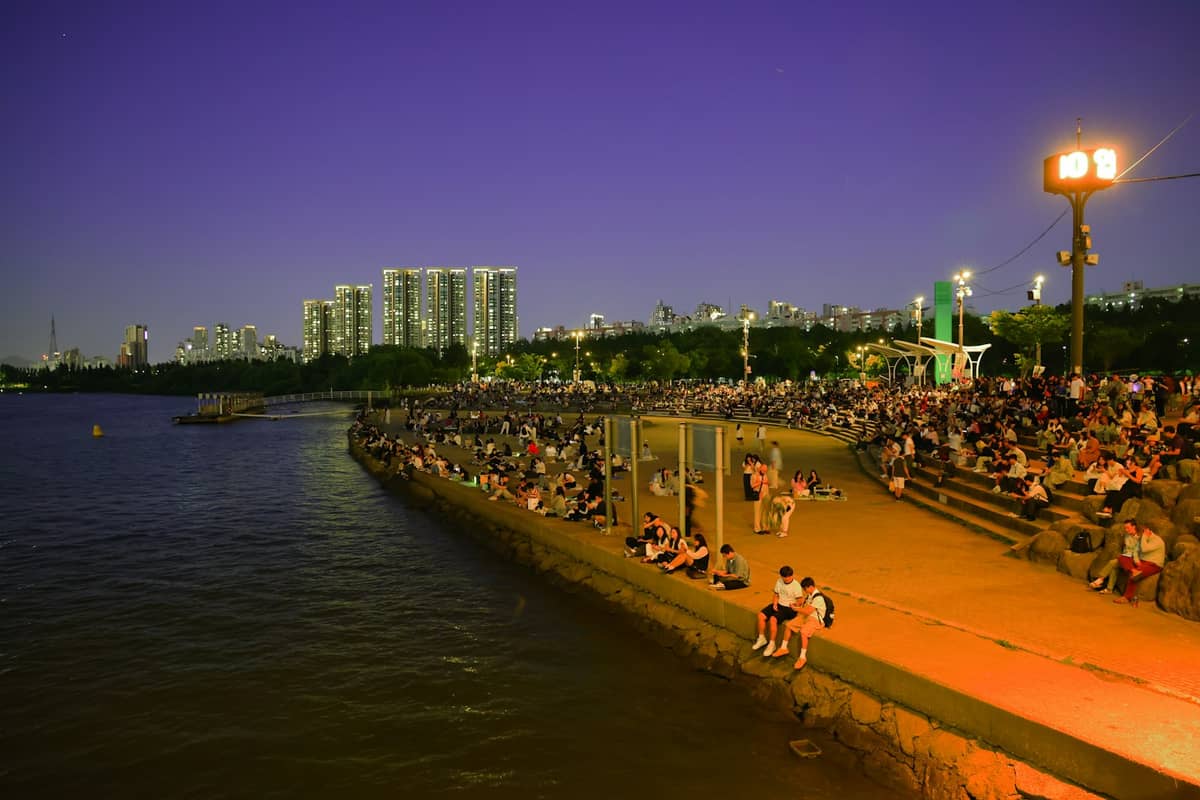 Multidão sentada ao longo da orla pavimentada do Rio Han no Parque Hangang de Yeouido, em Seul, ao entardecer, com o skyline da cidade e o céu lilás ao fundo.
