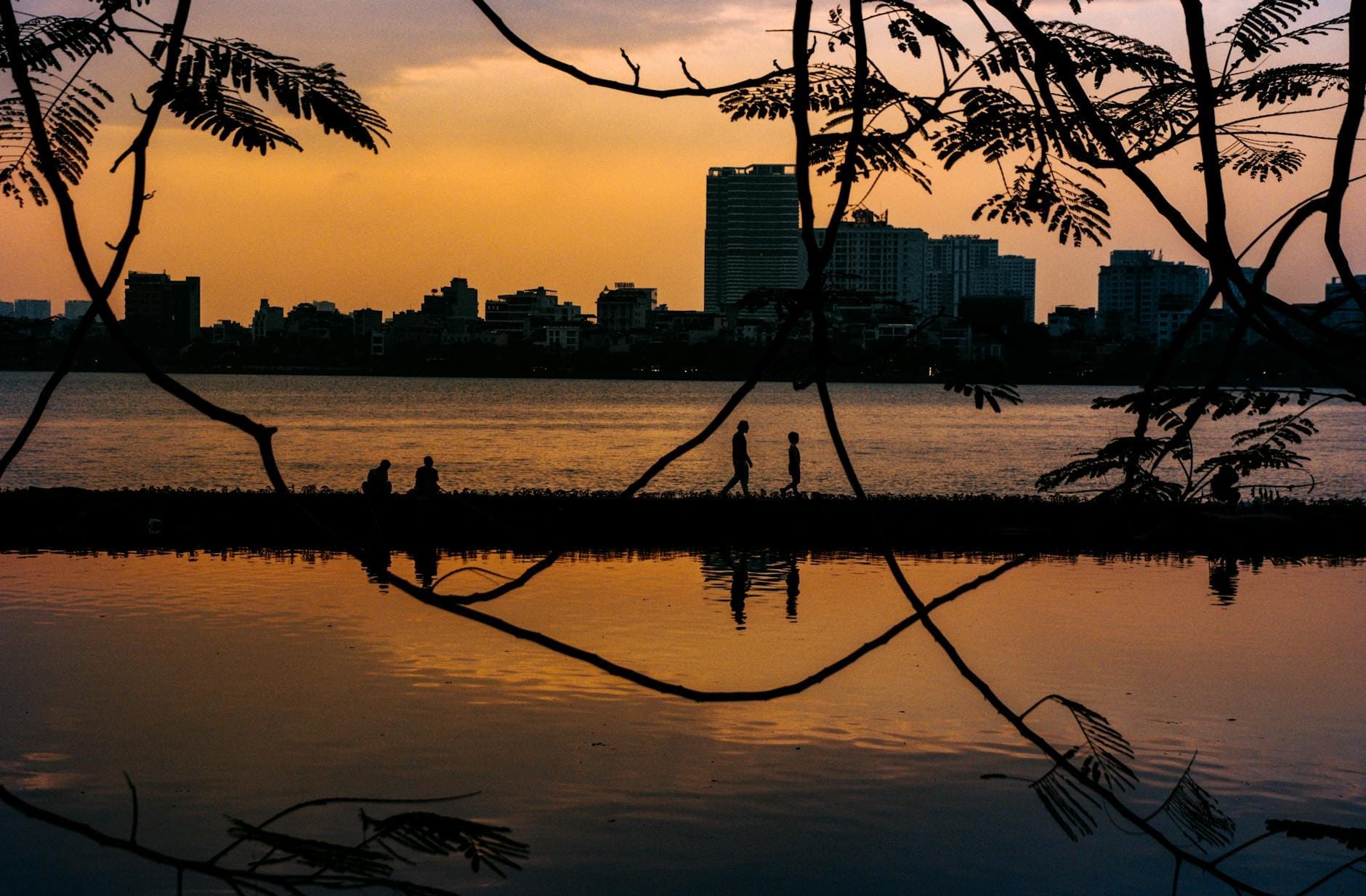 Silhouettes of people walking and sitting beside Hanoi's West Lake at sunset, with city buildings in the background.