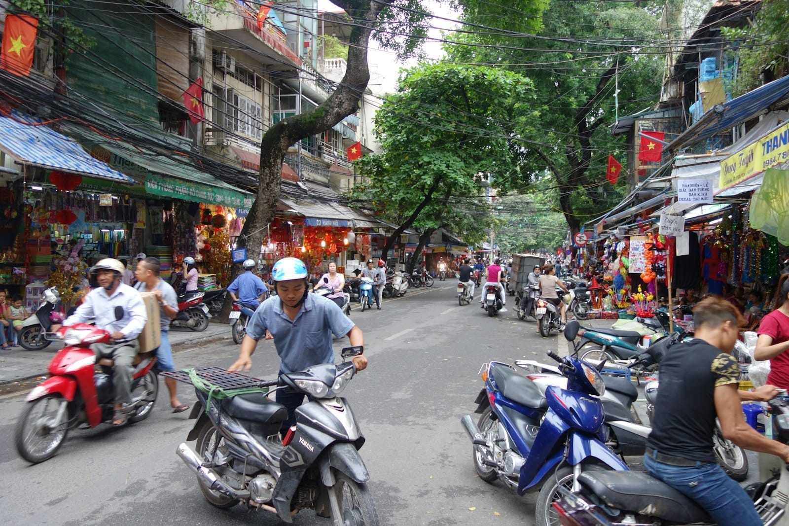 Bustling street in Hanoi Old Quarter with motorbikes, pedestrians, colorful shopfronts, and trees arching over the lively urban scene.