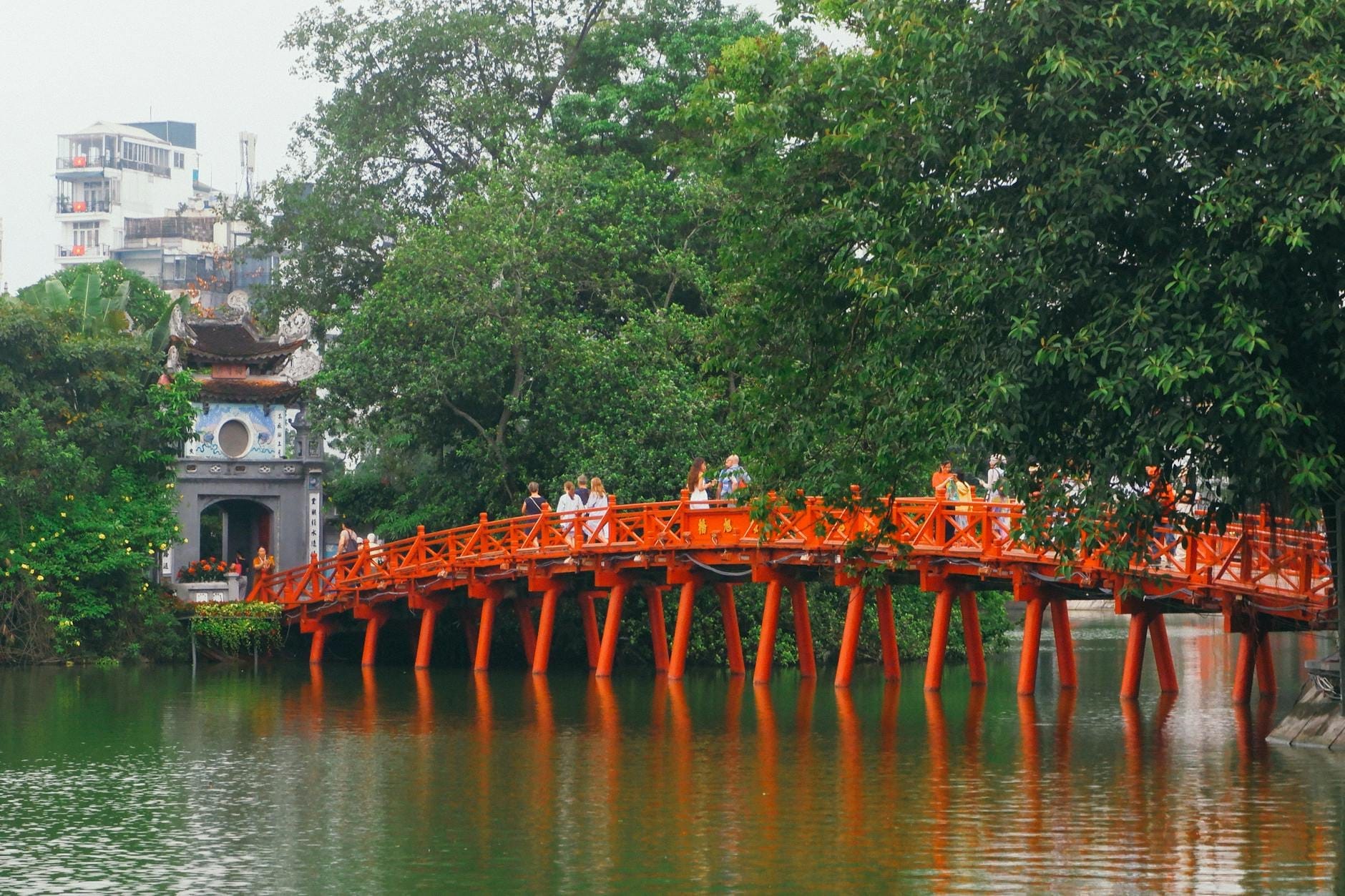 The iconic red Huc Bridge with people crossing, leading to the island temple on Hoan Kiem Lake in Hanoi Old Quarter.