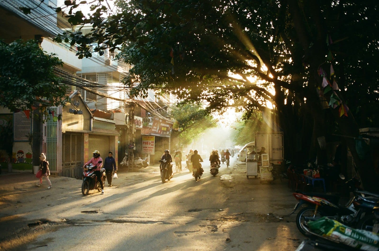 Morning sunlight streams through leafy trees onto a Hanoi Old Quarter street, with people riding motorbikes and walking along the lively road.