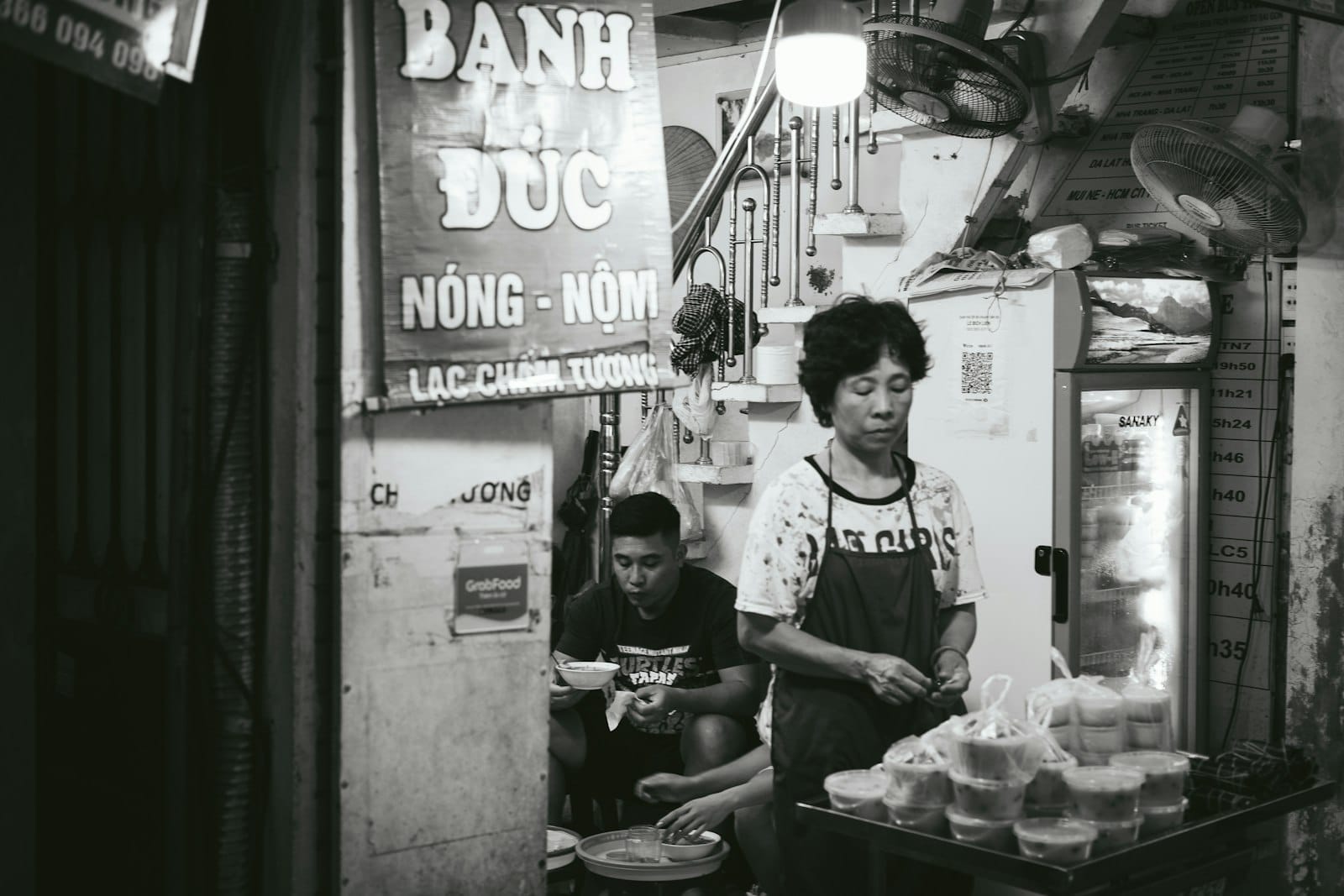 A woman preparing street food at a small stall in Hanoi Old Quarter, with steaming dishes and a traditional sign overhead.