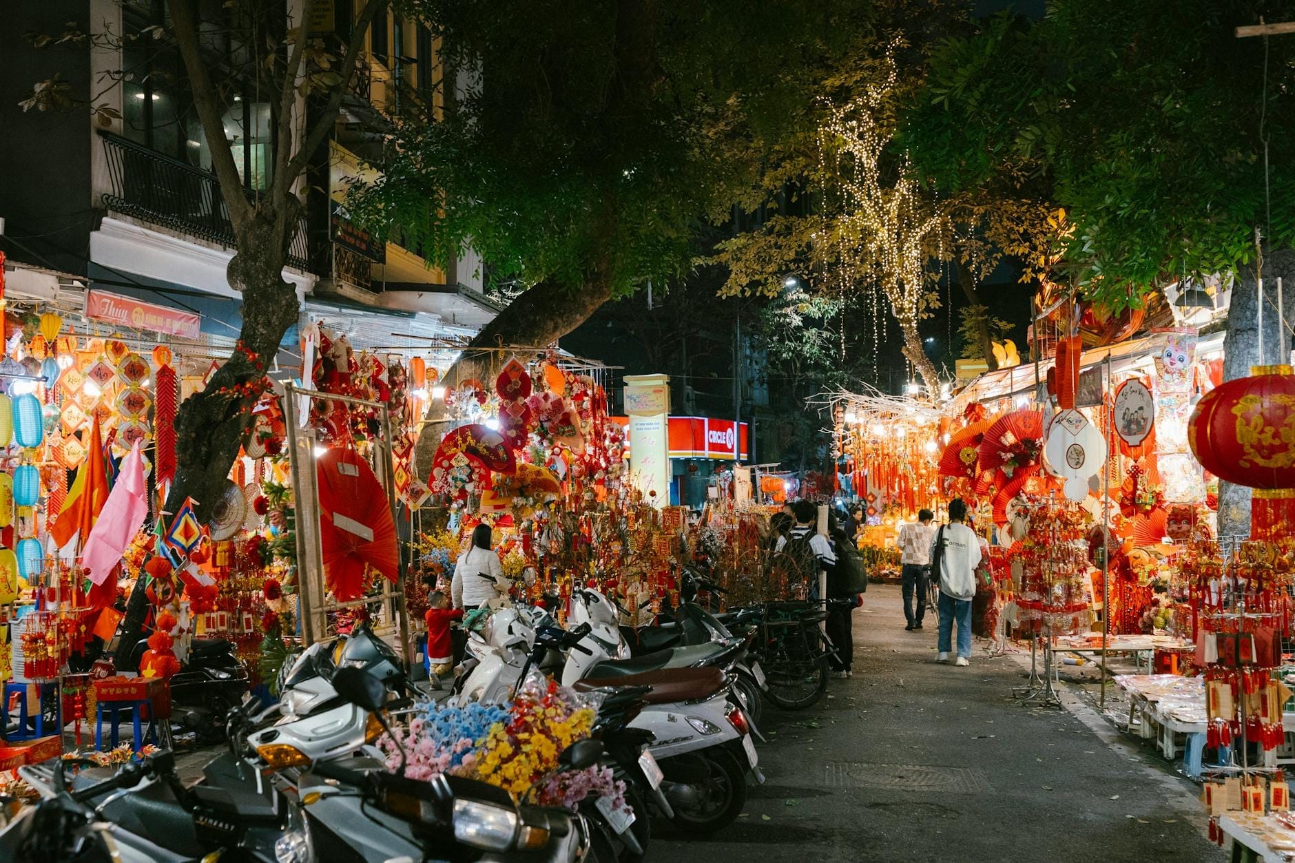 Night scene in Hanoi Old Quarter with market stalls, colorful lanterns, street decorations, and people walking among parked motorbikes under illuminated trees.