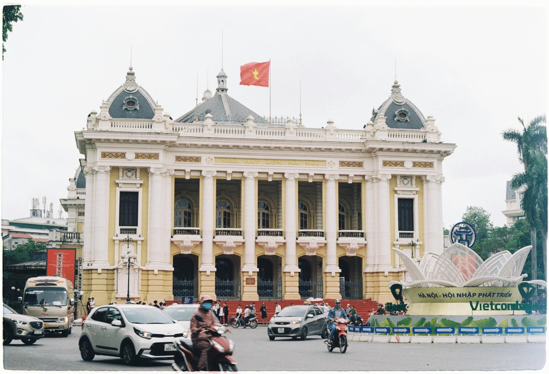 Daytime view of the Hanoi Opera House with a Vietnamese flag on top, showing the full neoclassical facade, bustling traffic, and people in front.