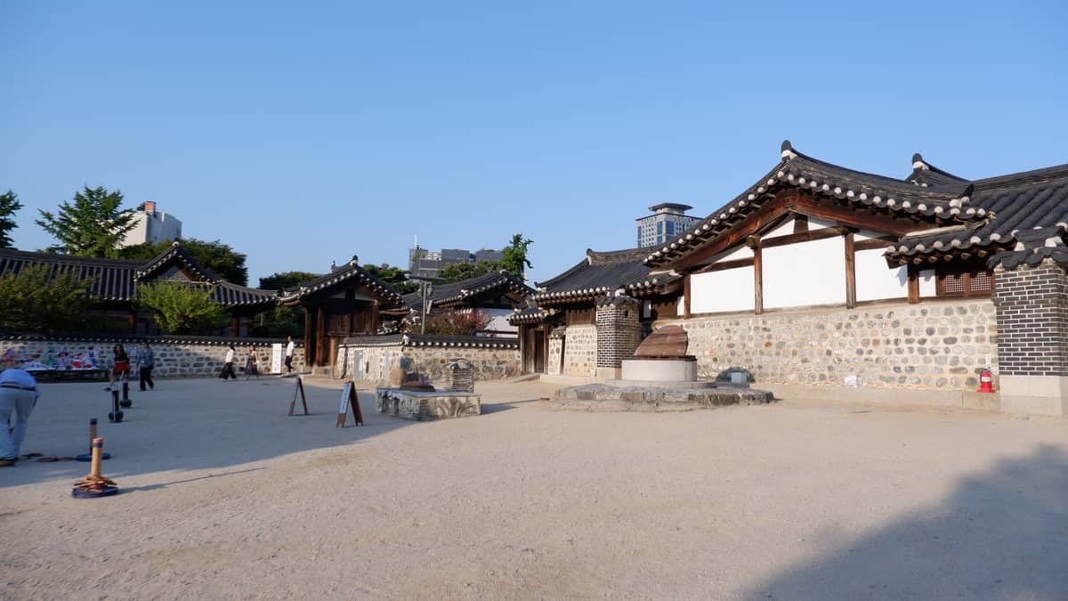 Pátio aberto com várias casas hanok tradicionais restauradas, chão de terra batida e edifícios da cidade ao fundo, provavelmente parte do Namsangol Hanok Village.