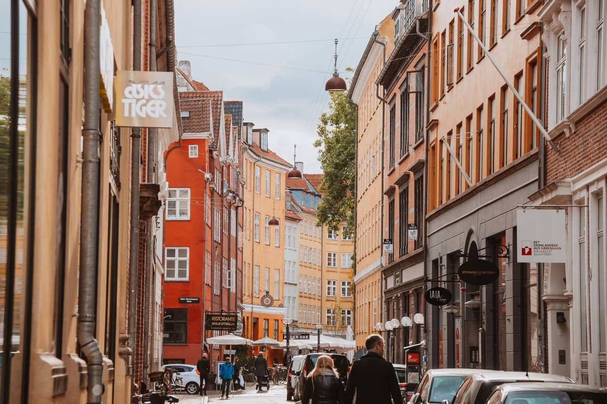 Un'incantevole strada di Copenaghen fiancheggiata da colorati edifici storici, persone che camminano, negozi e caffè sotto un cielo nuvoloso.