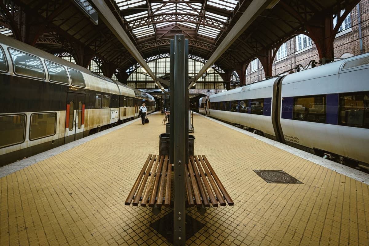 Binario della Stazione Centrale di Copenaghen con panchine, passeggeri e tipici treni danesi sotto un grande tetto ad arco.