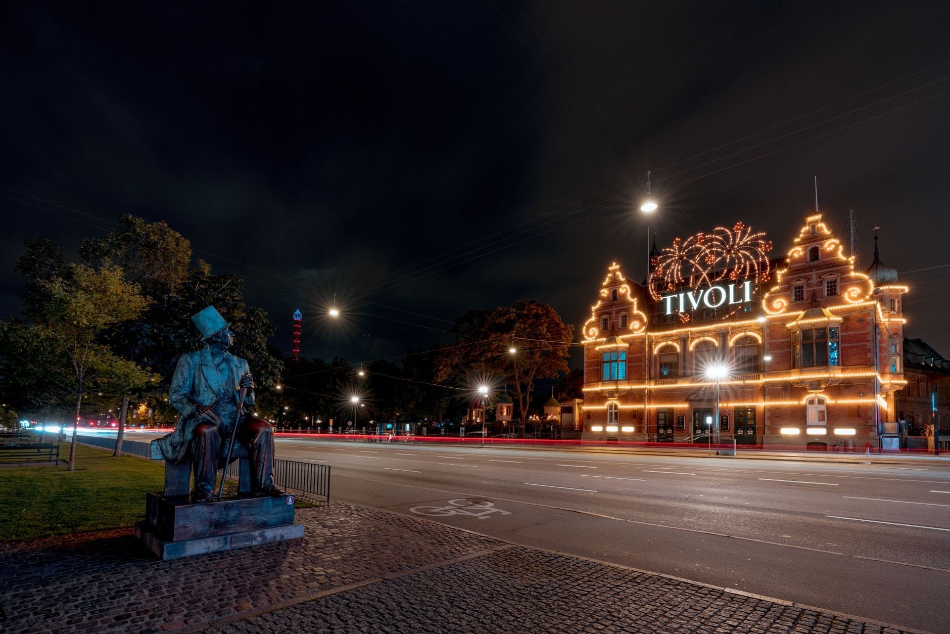 Cena noturna da entrada do Tivoli Gardens em Copenhague, lindamente iluminada, com uma estátua de bronze de Hans Christian Andersen em primeiro plano contemplando a cidade.