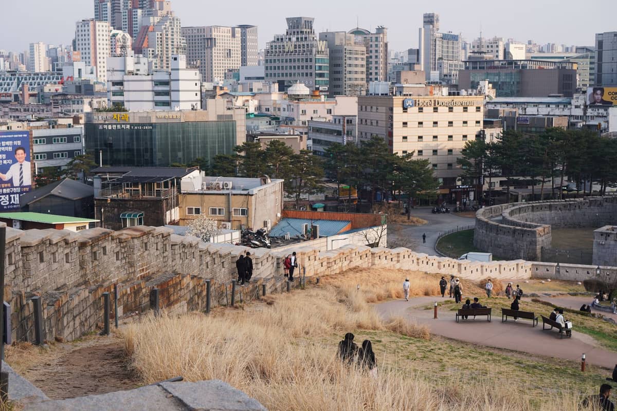 Vista da Hanyangdoseong (Muralha de Seul) com pessoas caminhando sobre as ameias de pedra e prédios urbanos ao fundo.