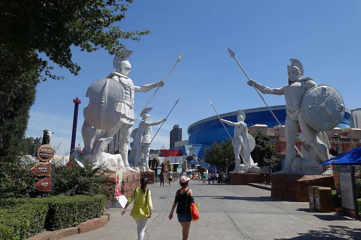 Riesige griechische Kriegerstatuen mit Schilden und Speeren flankieren den Haupteingangsweg des Happy Valley Beijing unter einem klaren blauen Himmel.