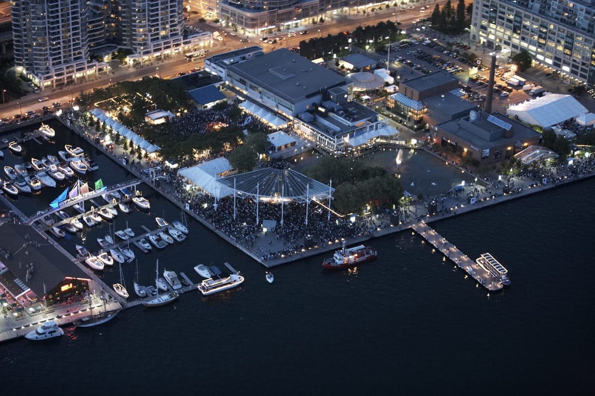 มุมมองทางอากาศของ Harbourfront Centre ในเมือง Toronto ยามพลบค่ำ แสดงให้เห็นอาคารริมน้ำ เต็นท์จัดงาน ท่าจอดเรือพร้อมเรือหลากหลายลำ และฝูงชนที่สนุกสนานในยามเย็น
